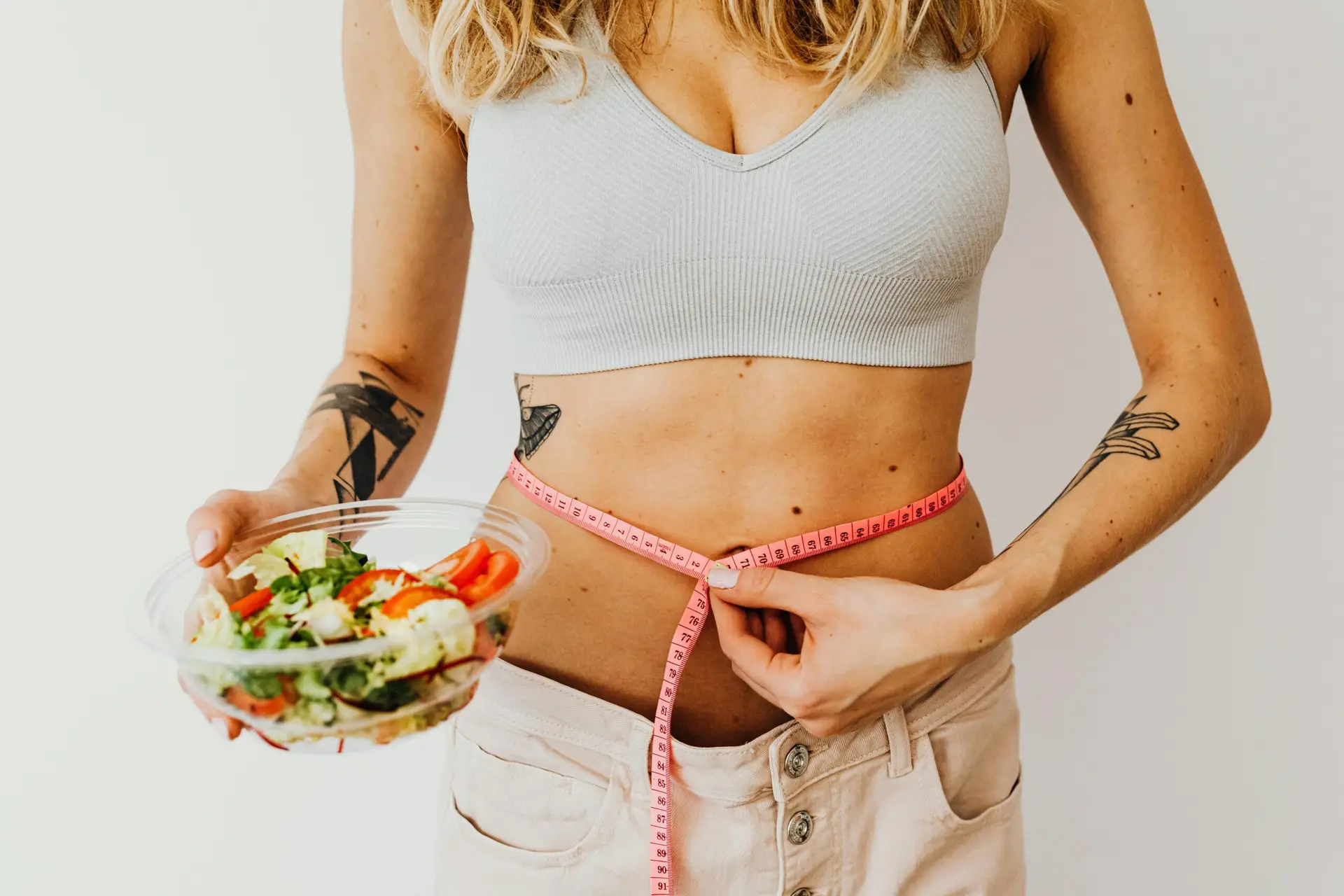 a woman holding in one hand a bowl of a a mixture of fruits and vegetables and with the other hand a tape measure around her waist and putting on loose pants to depict weight loss and things you need to do to have sustainable weight loss