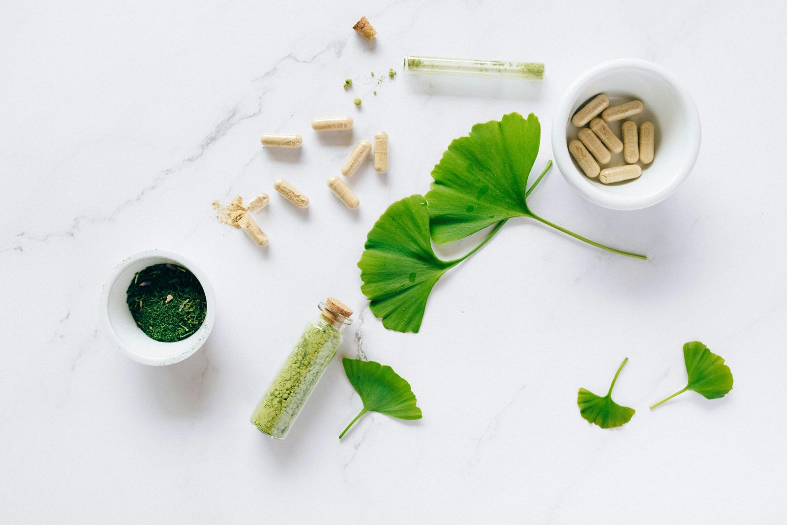 Image of a number of weight loss supplements laid out on a white background. there is a beautiful small white bowl of green tea extracts, there is another medium sized beautiful white bowl of weight loss capsules, there are also other capsules laid out. there are green leaves laid out on the background. there is also a small glass with dried out powder of green leaves.